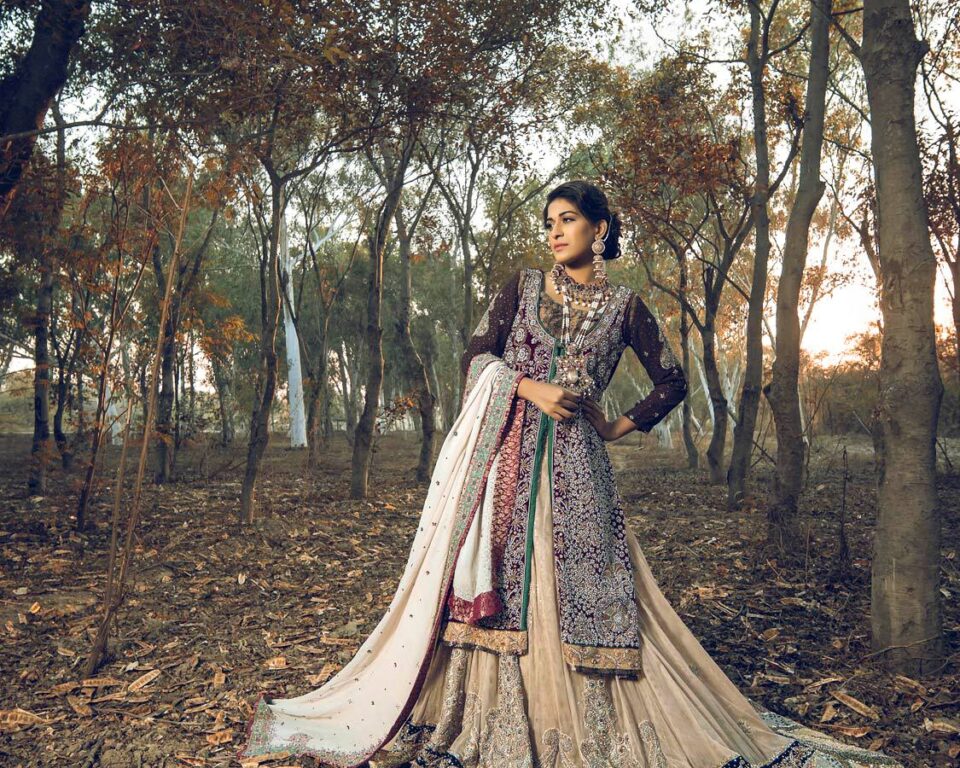 A woman in an elaborately embroidered, flowing traditional dress with shades of purple, gold, and cream stands in a sparse forest with dry leaves on the ground and trees with autumnal foliage, showcasing editorial fashion photography by Ahsan Ali Shahid