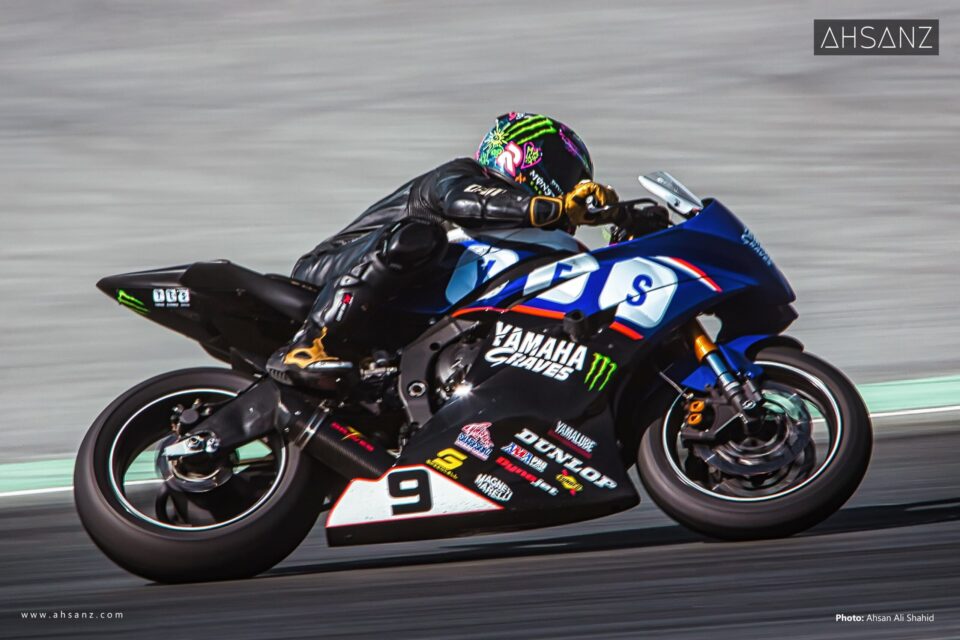 A motorcyclist in a black racing suit and a brightly colored helmet, riding a dark blue, black, and white Yamaha Graves racing motorcycle with the number 9, speeds around a turn on a grey racetrack.