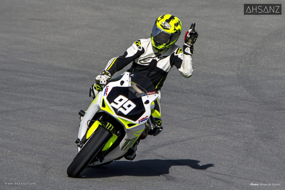 A motorcyclist in a white, black, and neon yellow racing suit and helmet, riding a white and neon yellow Honda CBR motorcycle with the number 99, leans into a turn on a racetrack, giving a thumbs up.