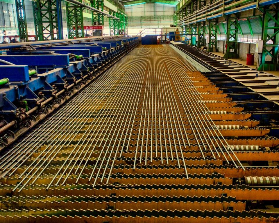 Wide-angle, high-dynamic-range photograph of a brightly lit factory floor, showing dozens of continuous steel wires or rebar being processed through industrial machinery with green and blue frames. Industrial photography by AHSANZ.