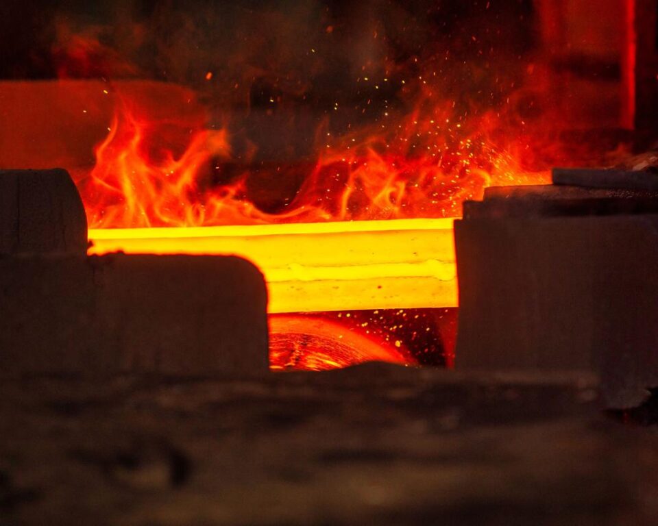 Extreme close-up photograph of a red-hot steel billet or bar glowing intensely orange-yellow with actual flames and sparks visible above it, emphasizing the immense heat of the industrial process. Industrial photography by AHSANZ for Mughal Steel.