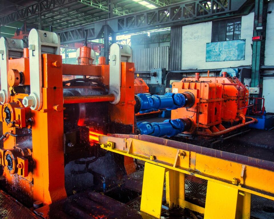 High-dynamic-range photograph inside a steel mill showing large orange and yellow rolling machinery processing glowing steel, with water cooling effects and steam. Industrial photography by AHSANZ for Mughal Steel.