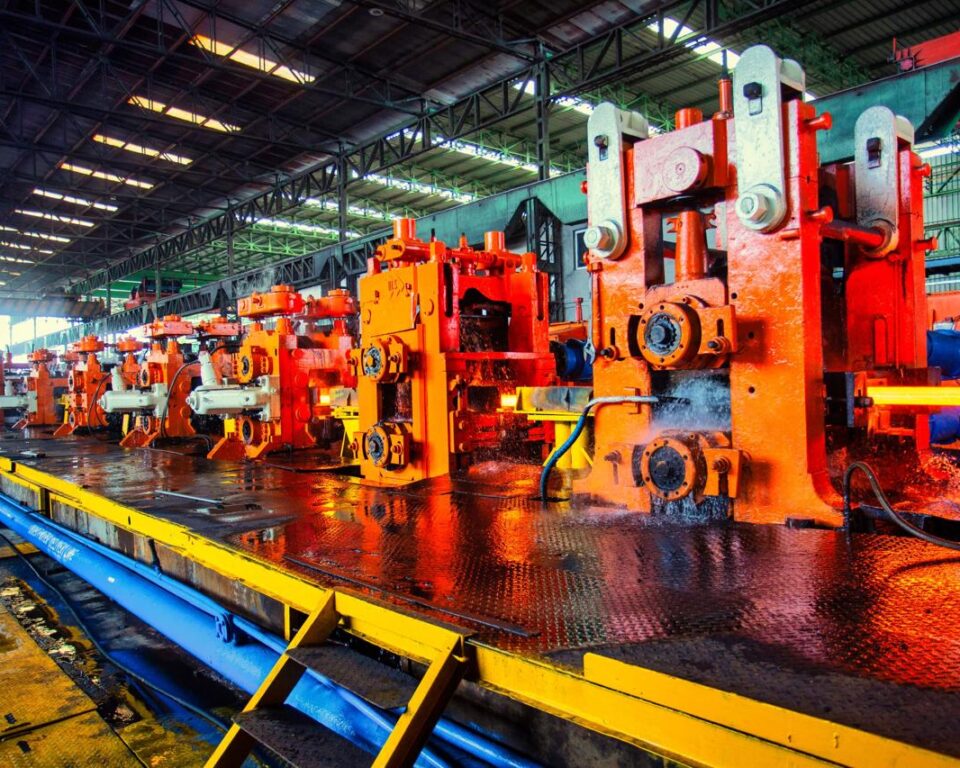 High-contrast photograph of a steel rolling mill interior, showing bright orange, red, and yellow industrial machinery processing hot metal with water cooling, emphasizing the dynamic atmosphere and heavy machinery. Industrial photography by AHSANZ for Mughal Steel.