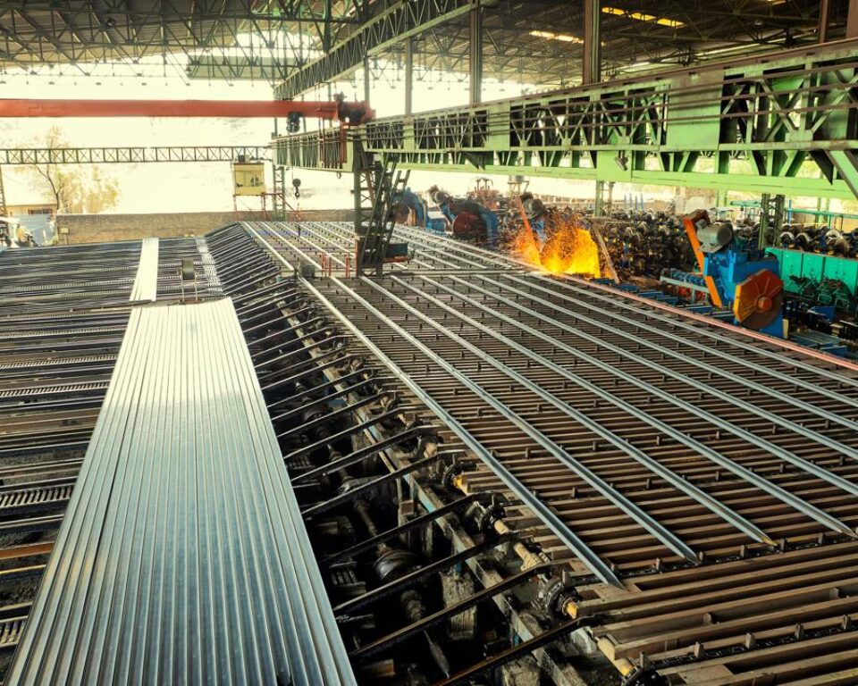 Wide-angle industrial photograph of a large steel mill facility, showing long metal sheets or rebar bars being processed on an active cooling bed, with workers and bright sparks visible in the background. Industrial photography by AHSANZ for Mughal Steel.