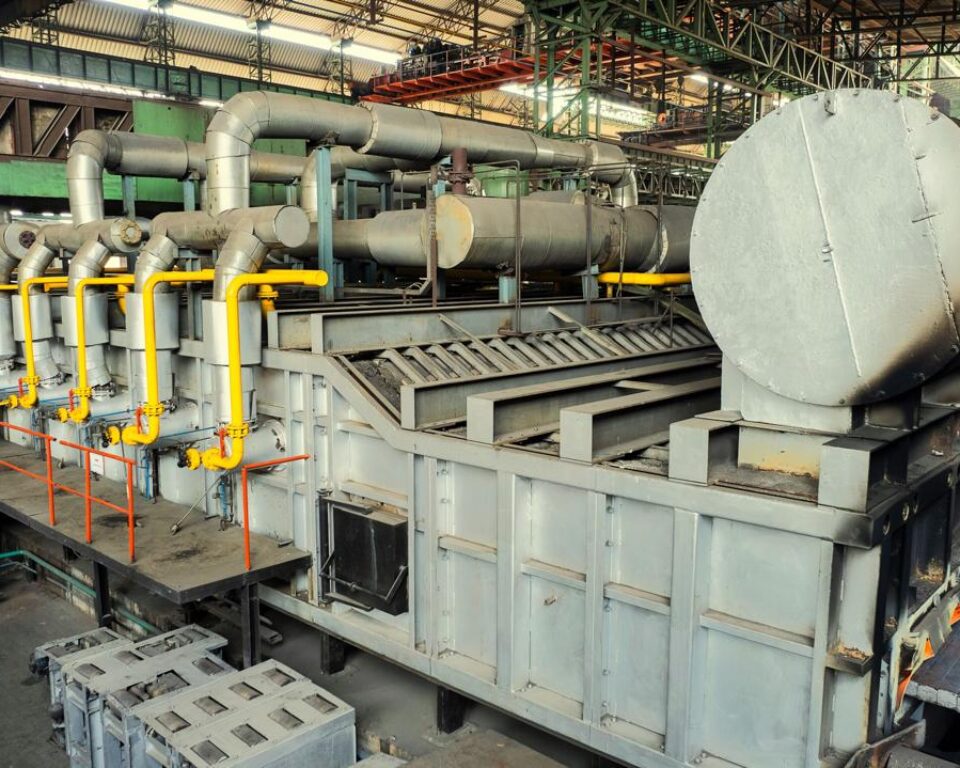 High-angle industrial photograph of a large, cylindrical, gray metal industrial unit inside a factory, with numerous yellow pipes and control valves running along the top structure. Industrial photography by AHSANZ for Mughal Steel.