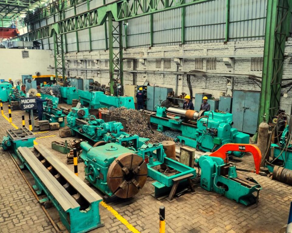 Wide-angle, interior photograph of a large machine shop floor, featuring numerous teal-colored heavy industrial lathes and machinery, with metal shavings piled in the foreground and factory workers visible. Industrial photography by AHSANZ for Mughal Steel.