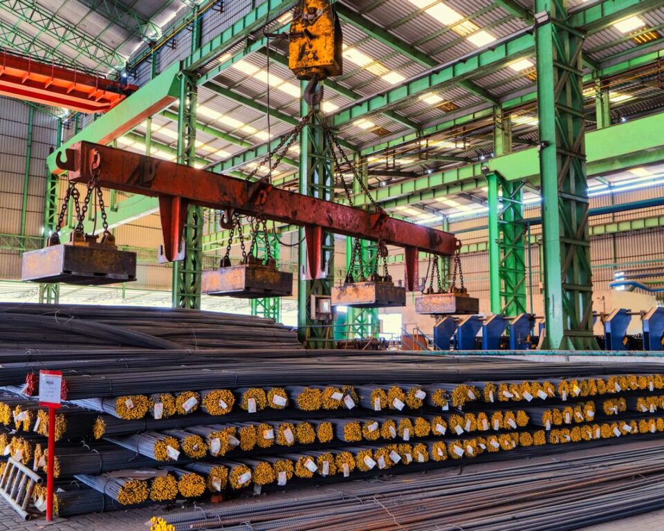 Close-up, high-dynamic-range photograph inside a steel mill showing red-hot, glowing steel billets or rebar moving along a dark, heavy-duty industrial conveyor belt, with ambient light. Industrial photography by AHSANZ for Mughal Steel.
