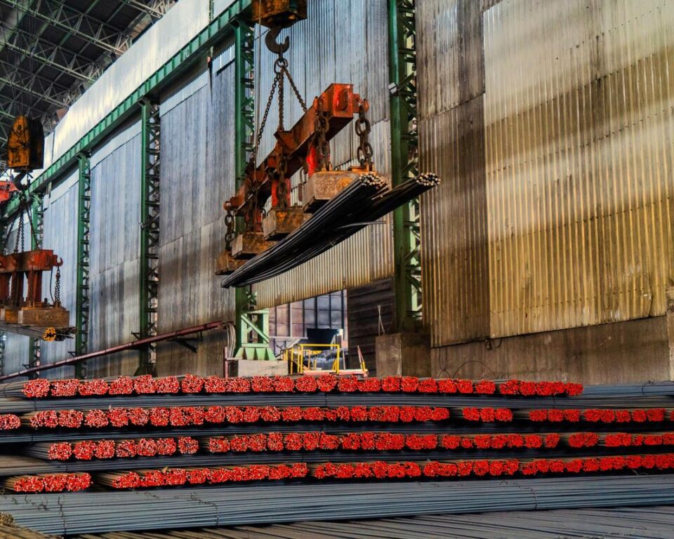 Wide-angle photograph inside a steel mill showing vast stacks of bundled rebar (reinforcing bars), with a heavy overhead crane using magnetic lifters to transport bundles of steel. Industrial photography by AHSANZ for Mughal Steel.